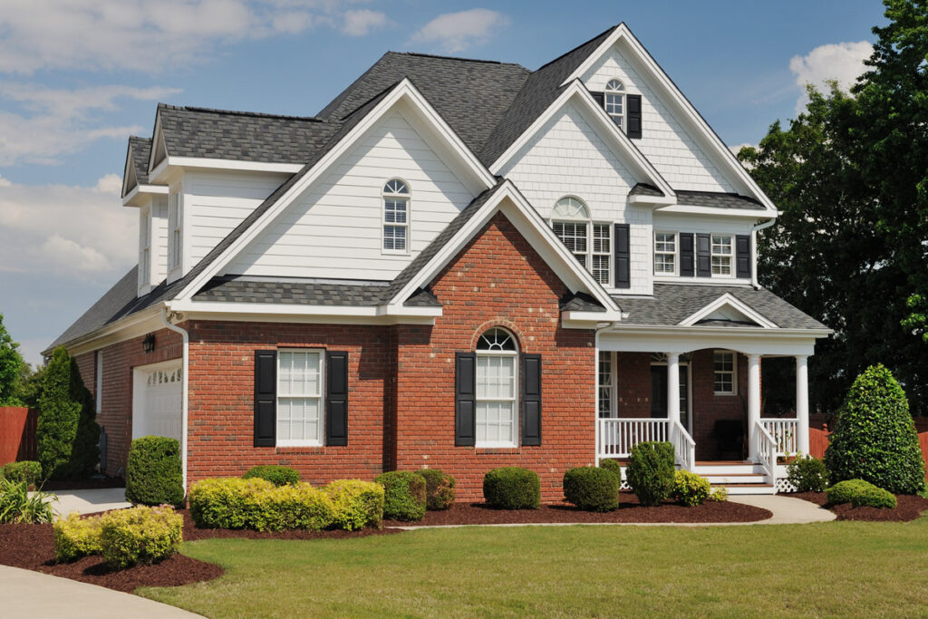 Two-story suburban house with red brick and white siding, front porch, black shutters, and landscaped lawn on a sunny day.