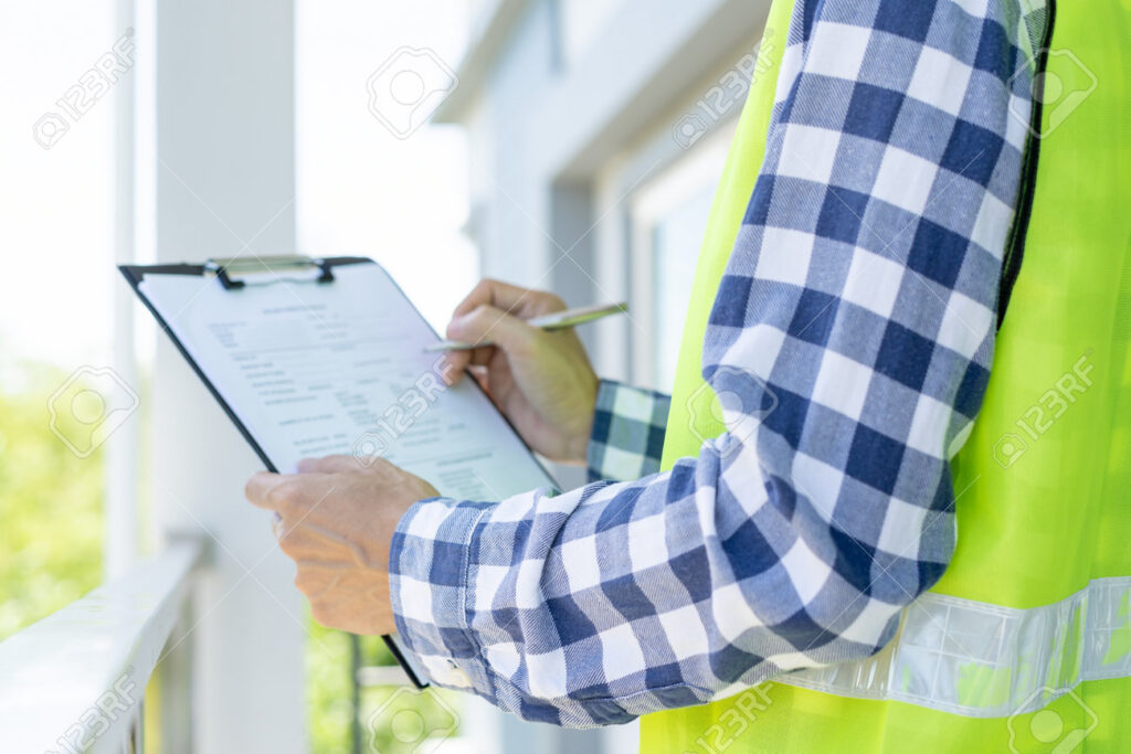 Person in a checked shirt and safety vest writing on a clipboard outdoors.